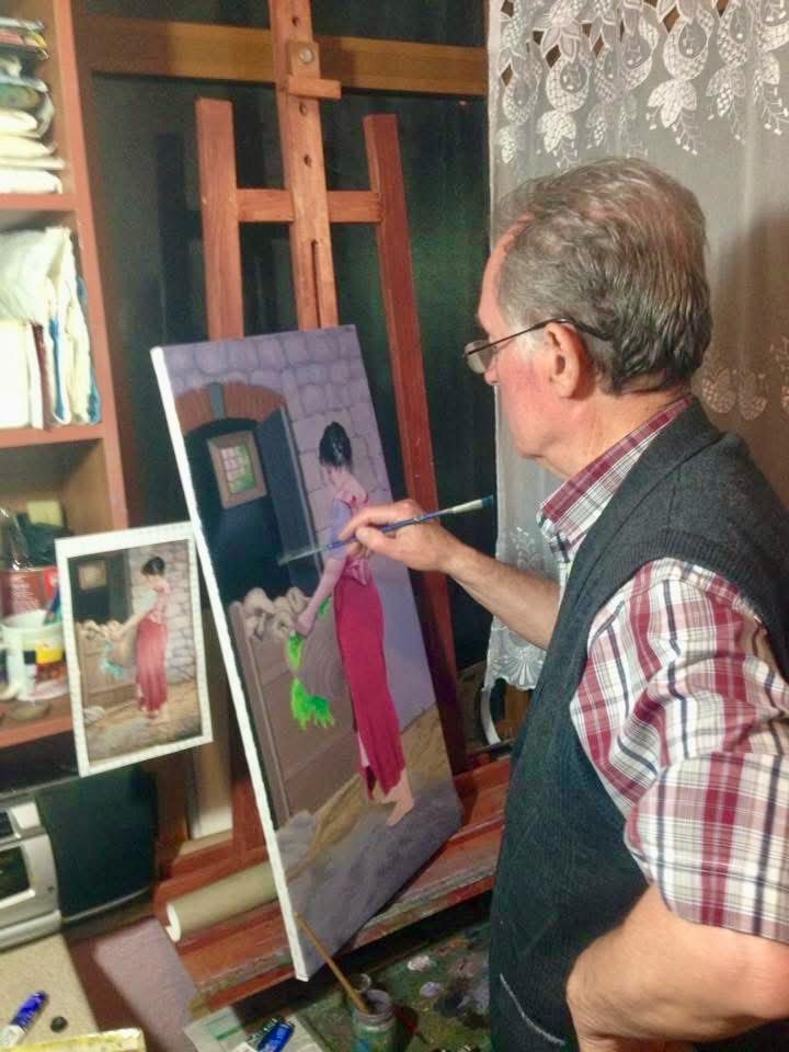 Gëzim Begolli holding a paintbrush at his easel, working on a painting of a woman at a fountain, with a reference photograph and art materials nearby.