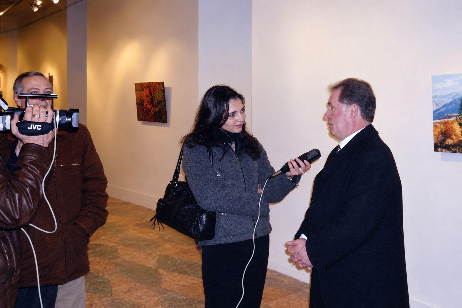 Gëzim Begolli speaking to an interviewer while a camera operator films, with paintings on the gallery walls.