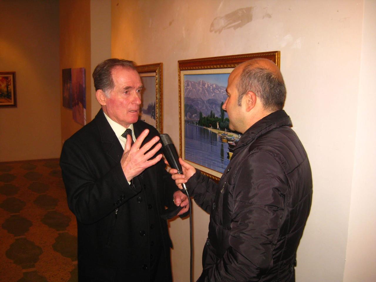 Gëzim Begolli speaking to a man holding a microphone in an exhibition gallery, beside framed paintings of mountains and boats.